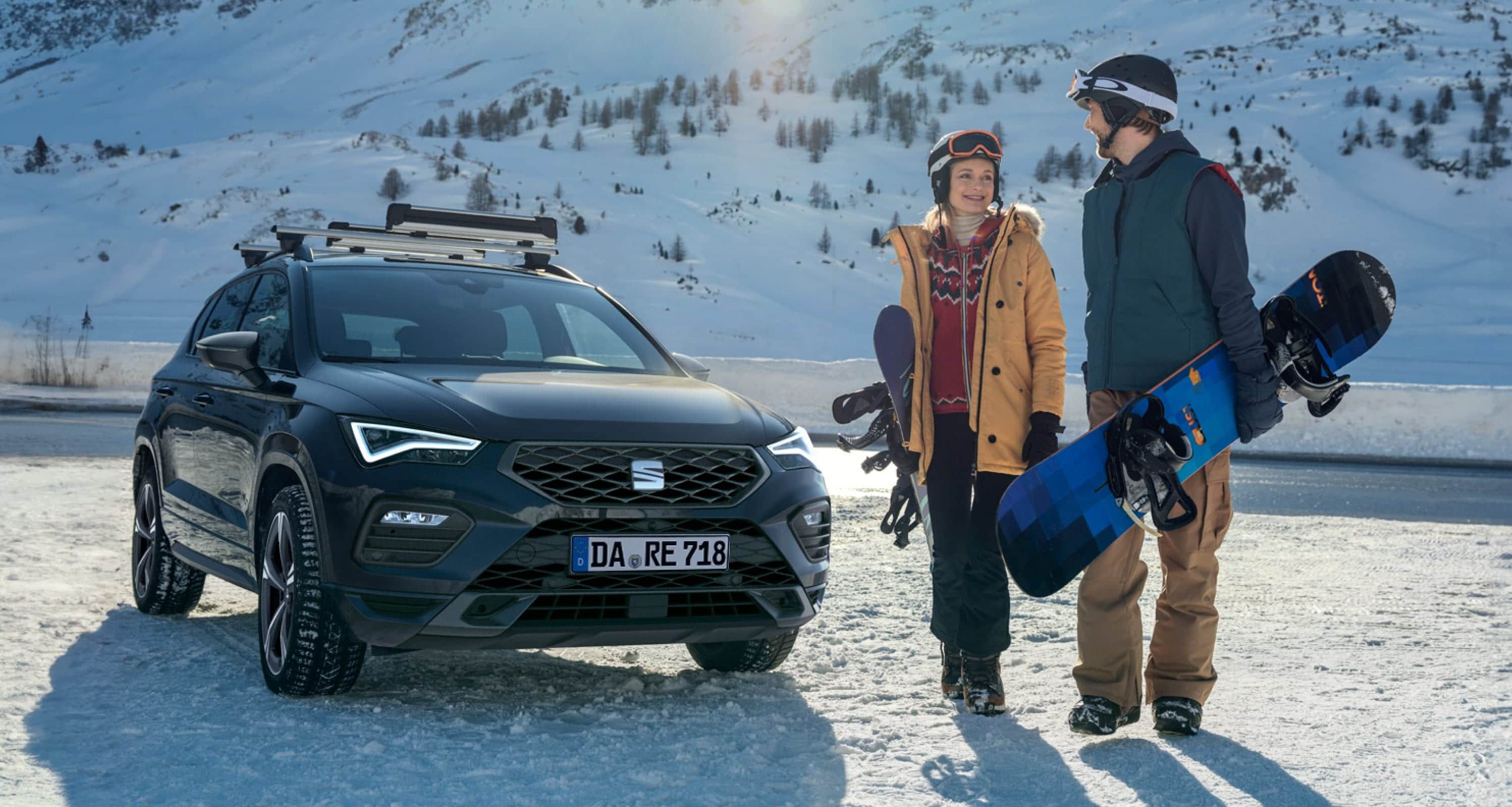 Ein EAT Ateca SUV parkt auf verschneitem Boden, daneben stehen zwei Personen mit Snowboards. Winterkompletträder 2024 von SEAT, ideal für verschneite Straßen.