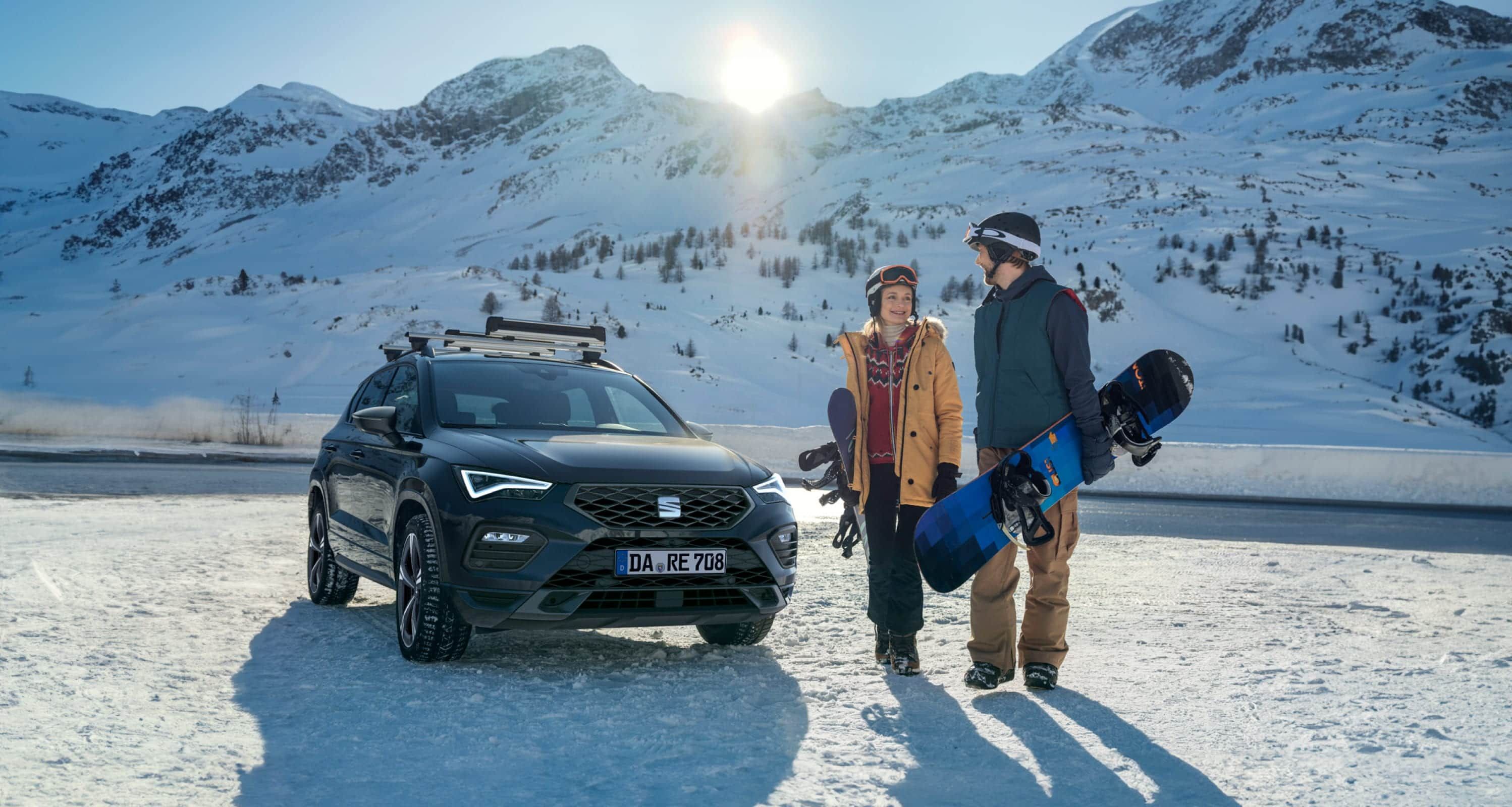 Ein EAT Ateca SUV parkt auf verschneitem Boden, daneben stehen zwei Personen mit Snowboards. Winterkompletträder 2024 von SEAT, ideal für verschneite Straßen.