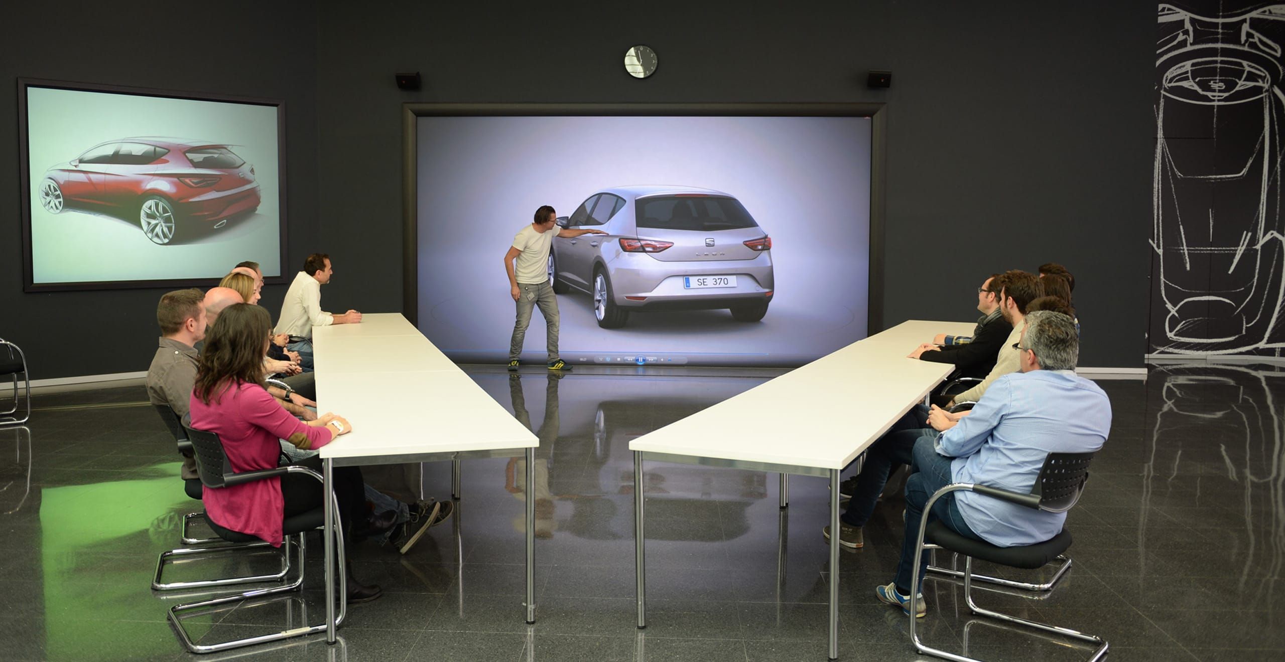 Gesprächsrunde ein Mann zeigt auf ein Autodesign SEAT Leon mit einer Gruppe von Leuten an einem Tisch, die zusehen – SEAT Karriere